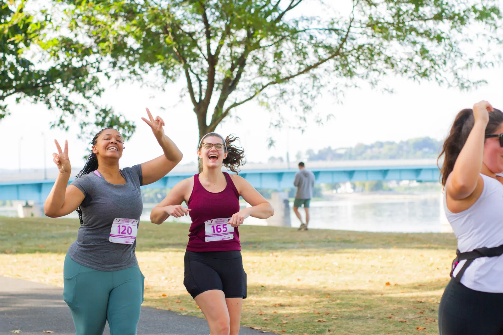 Two women smiling and giving peace signs while running a race, with a bridge and river in the background.