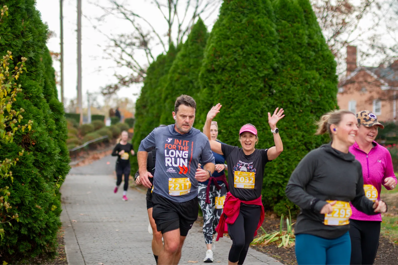 Runners smiling and waving as they participate in a race, with trees lining the course.