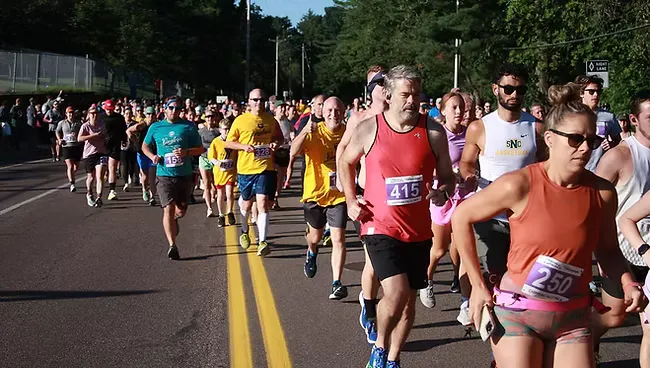 A large group of runners are starting a race on a paved road, with trees and foliage in the background.