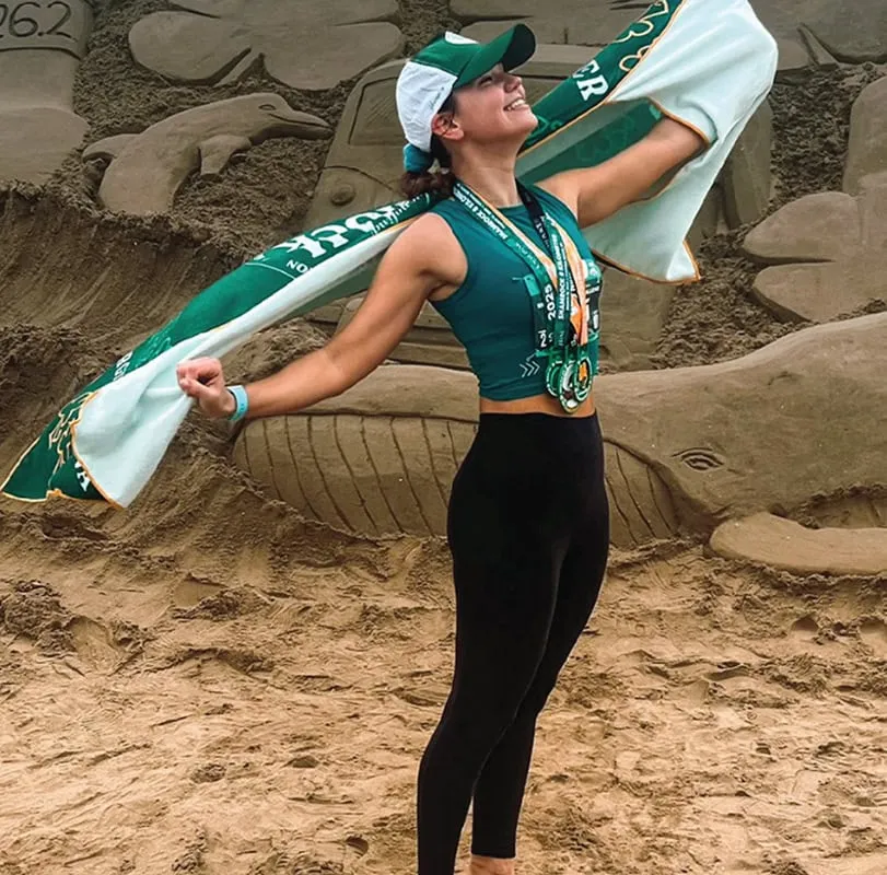 A runner celebrates with medals and a race scarf in front of a sand sculpture on a beach.