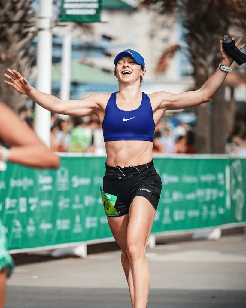 A female runner with arms outstretched in celebration, wearing a blue sports bra and black shorts, crossing the finish line of a race.