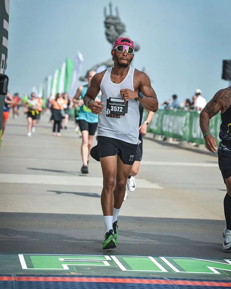 A male runner wearing a white tank top, black shorts, and a pink bandana crosses the finish line of a race.