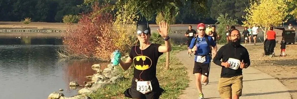 A runner dressed as Batman waves while participating in a race near a lake with fall foliage.