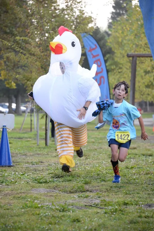 A child runner with bib number 1422 runs alongside a person in a large inflatable chicken costume at the start of a race.