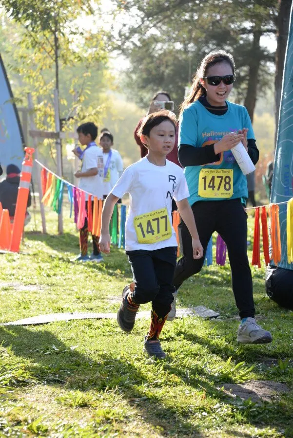Two young runners, one in front wearing a white shirt with bib number 1477, and another slightly behind wearing a teal shirt with bib number 1478, running on a grassy course with colorful ribbons marking the path.